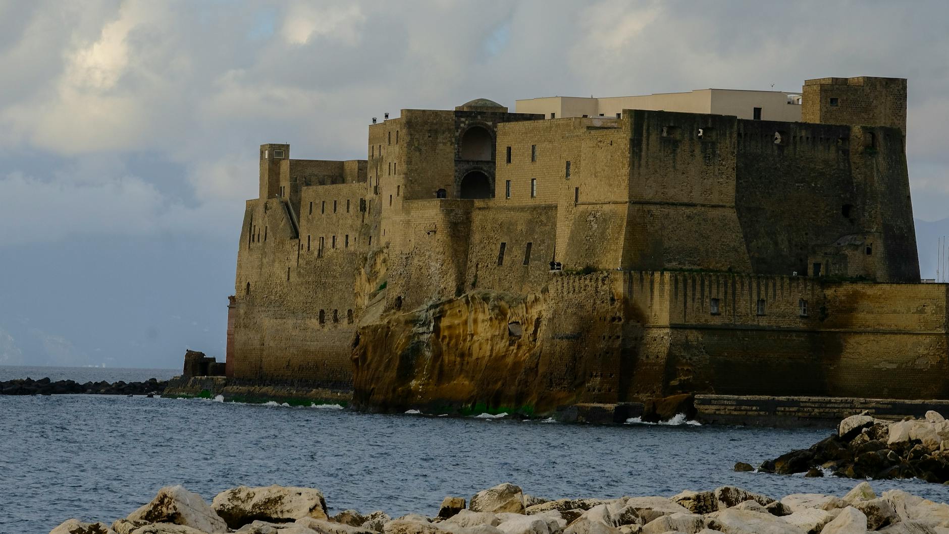 Scenic view of Castel dell'Ovo in Naples, Italy, with rocky shore and clouds.