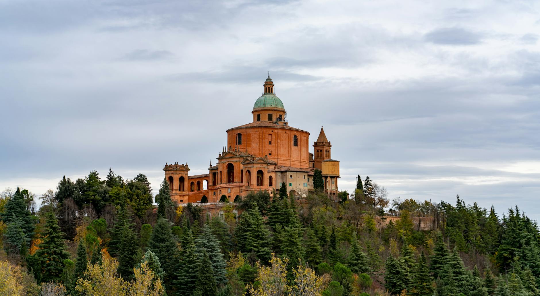 The historic Sanctuary of Madonna di San Luca sits atop a lush hill with a dramatic sky.