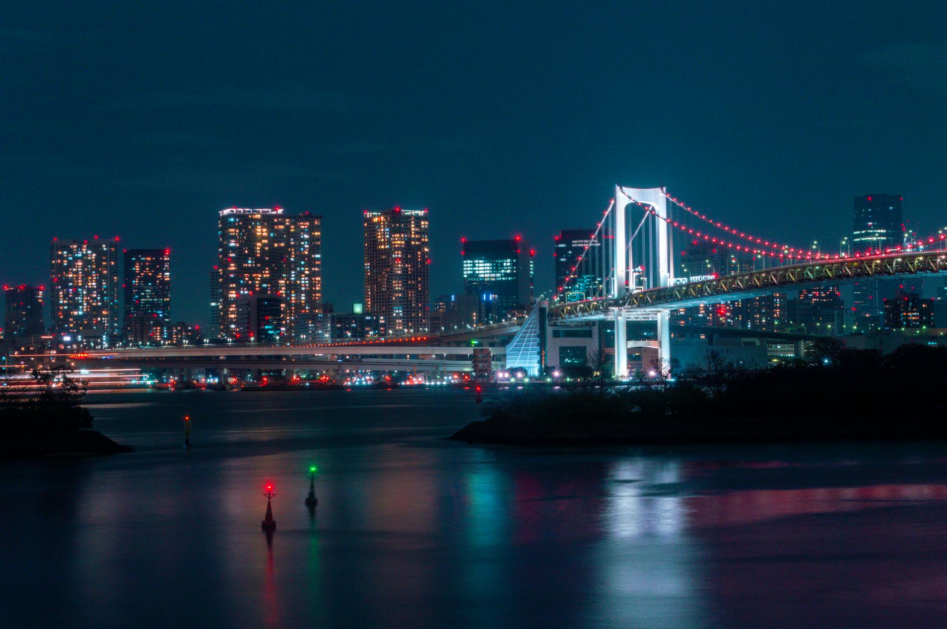 Night view of a brightly lit bridge and city skyline reflected on a calm river.