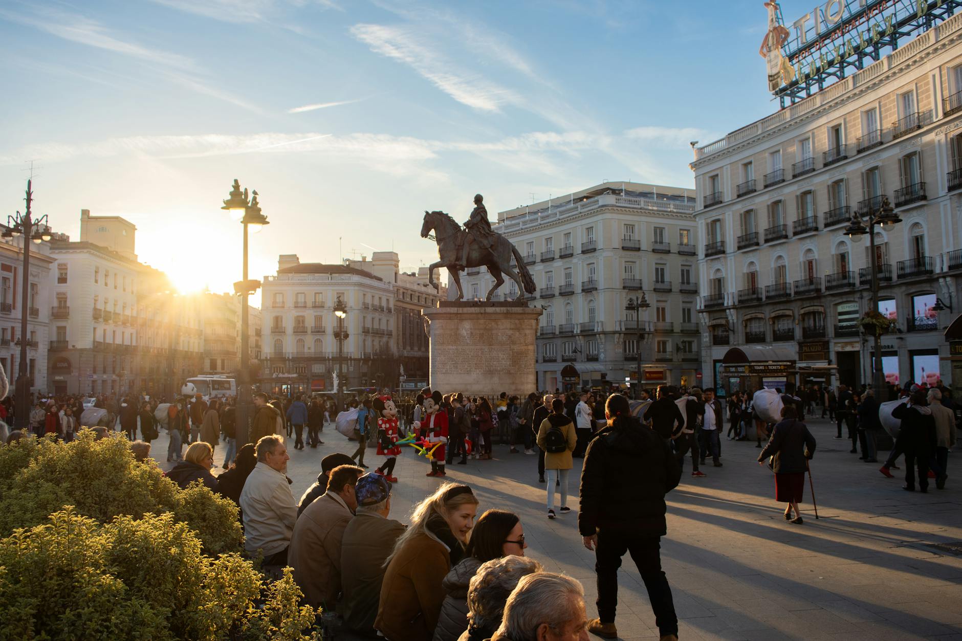 Tourists and locals gather at Puerta del Sol in Madrid during sunset, with the statue of King Charles III in view.